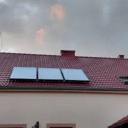 Trzy panele słoneczne zamontowane na czerwonym dachu domu jednorodzinnego, widoczne rynny i okno dachowe, pochmurne niebo w tle.