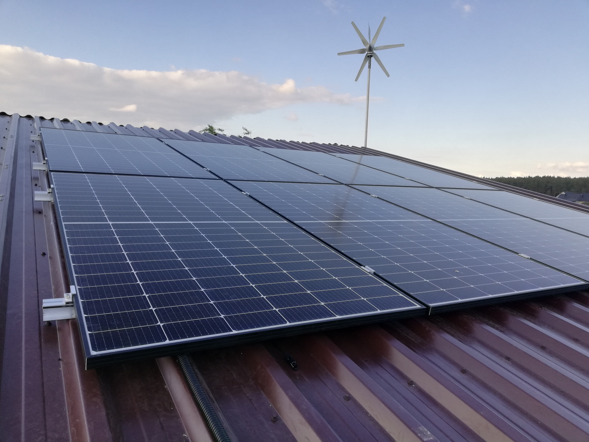 Panel słoneczny na brązowym dachu z turbiną wiatrową w tle, widok z góry, częściowo zachmurzone niebo.