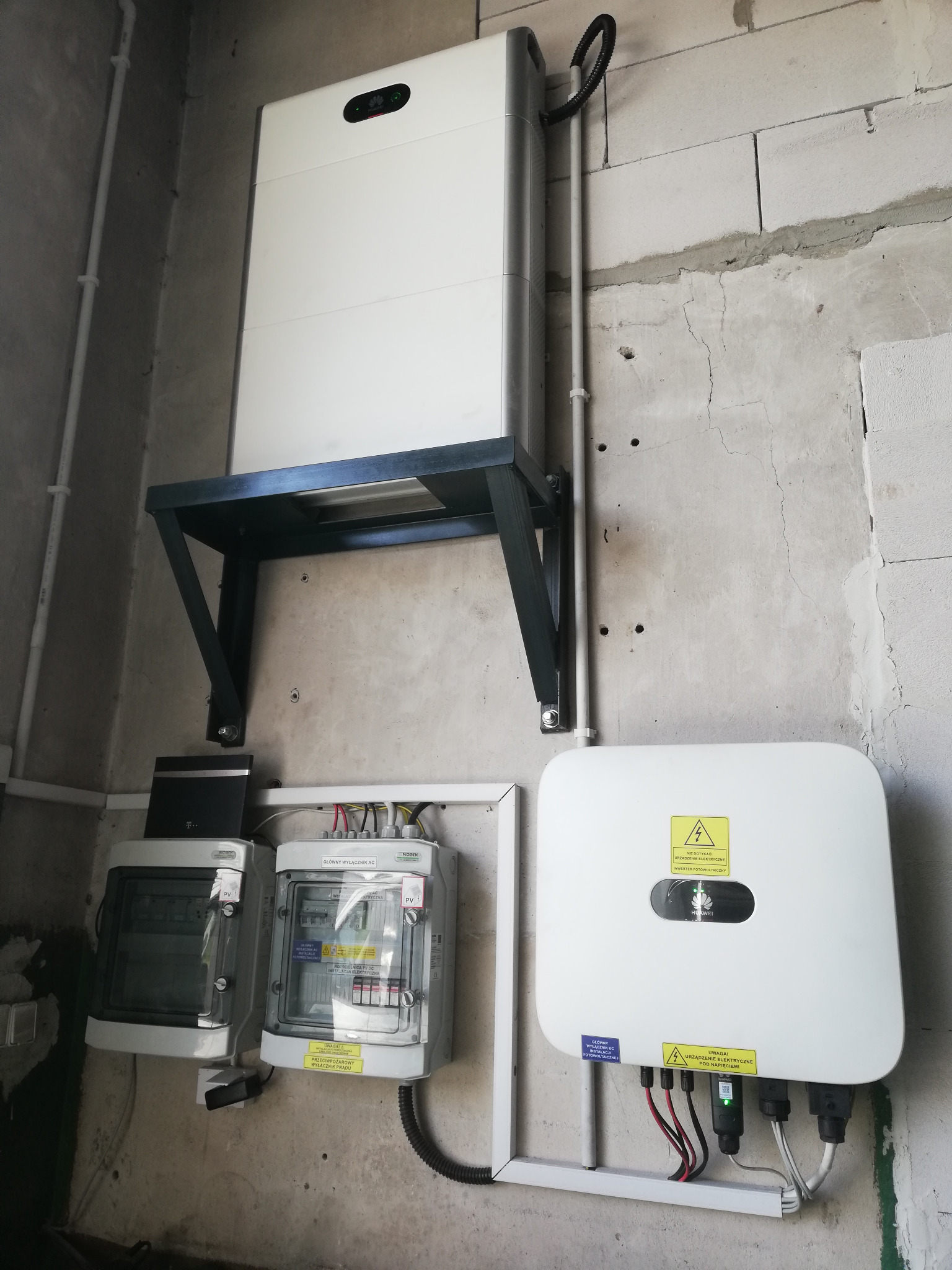Bateria i falownik instalacji fotowoltaicznej zamontowane na surowej ścianie z betonu komórkowego. Widoczne przewody, skrzynki z bezpiecznikami i elementy mocujące.