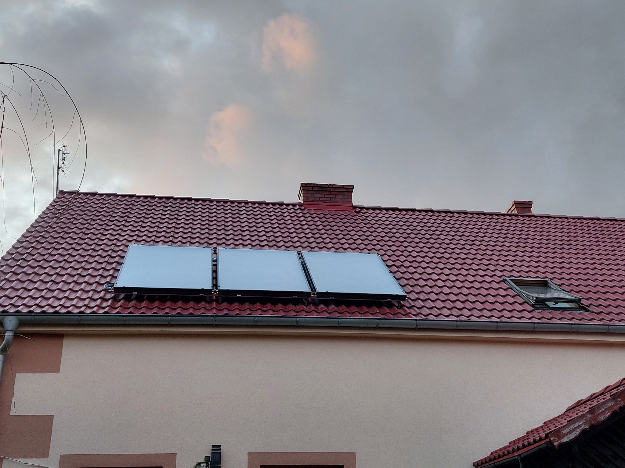Trzy panele słoneczne zamontowane na czerwonym dachu domu jednorodzinnego, widoczne rynny i okno dachowe, pochmurne niebo w tle.