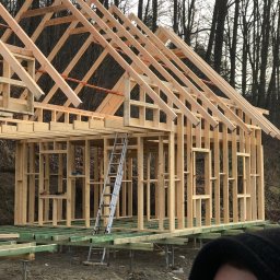 Altany tarasy - Surowa drewniana konstrukcja szkieletowa budynku w trakcie budowy, widoczna drabina oparta o konstrukcję, tło stanowią nagie drzewa.