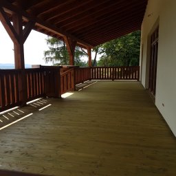 Altany tarasy - Drewniany taras z balustradą i zadaszeniem, widok na góry w tle, promienie słońca padające na deski podłogowe.