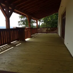 Drewniany taras z balustradą i zadaszeniem, widok na g&oacute;ry w tle, promienie słońca padające na deski podłogowe.