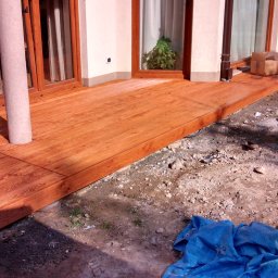 Altany tarasy - Nowy taras z desek kompozytowych w odcieniach brązu, z widocznym słupem podtrzymującym i fragmentem ogrodu w trakcie aranżacji.