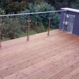 Altany tarasy - Nowoczesny taras z drewnianą podłogą i szklaną balustradą ze stalowymi słupkami, widok na zieleń w tle, folia ochronna na elemencie ściany.