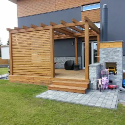 Drewniana pergola z ażurową ścianą boczną przylegająca do domu z kominkiem zewnętrznym, widok na taras z meblami ogrodowymi.