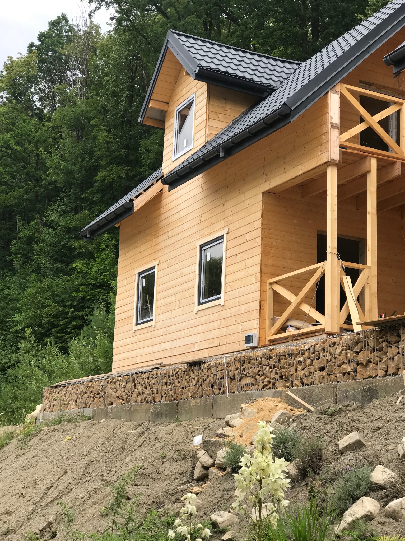 Surowa drewniana konstrukcja domu z szarą dachówką, widoczne okna z białymi ramami, zabezpieczenie terenu gabionami wypełnionymi kamieniami, w tle zieleń drzew.