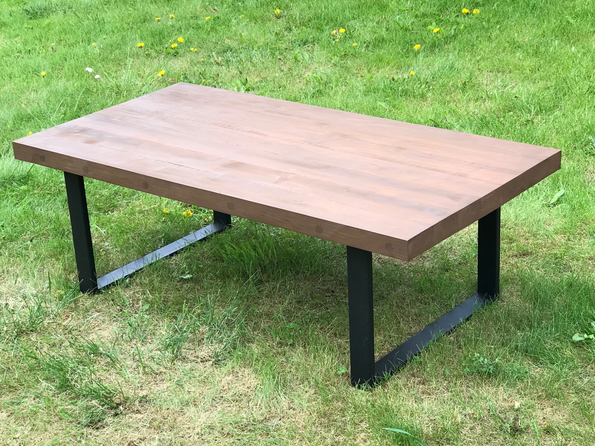 Prostokątny stolik kawowy z drewnianym blatem w odcieniach brązu i czarnymi metalowymi nogami, ustawiony na trawniku z żółtymi kwiatami.