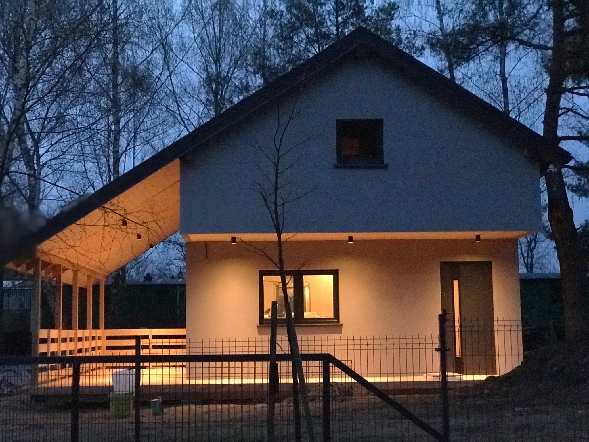 Nowoczesny dom z dobudowaną, oświetloną konstrukcją z drewna, widoczny zza metalowego ogrodzenia w szarówce.