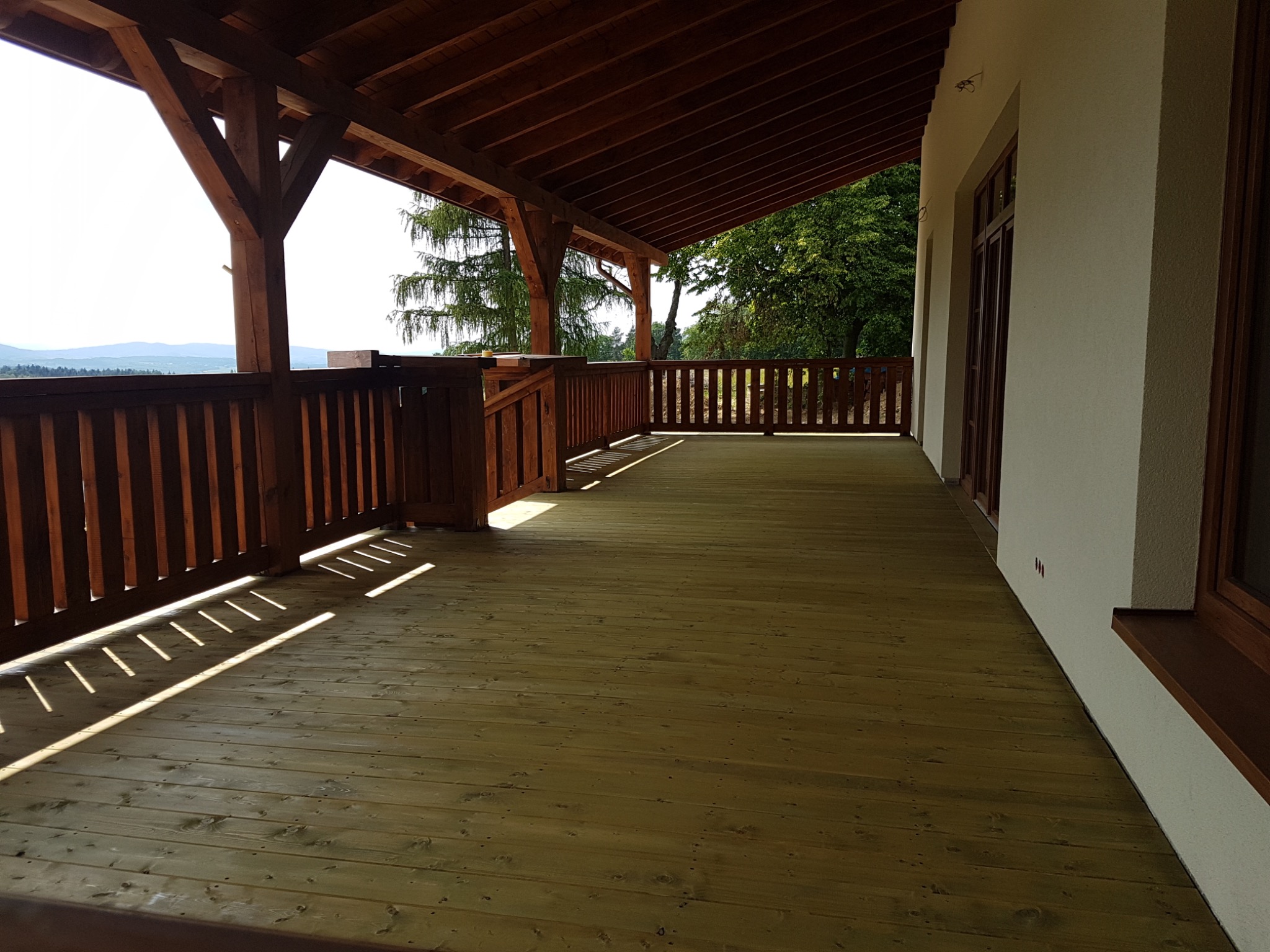 Drewniany taras z balustradą i zadaszeniem, widok na góry w tle, promienie słońca padające na deski podłogowe.