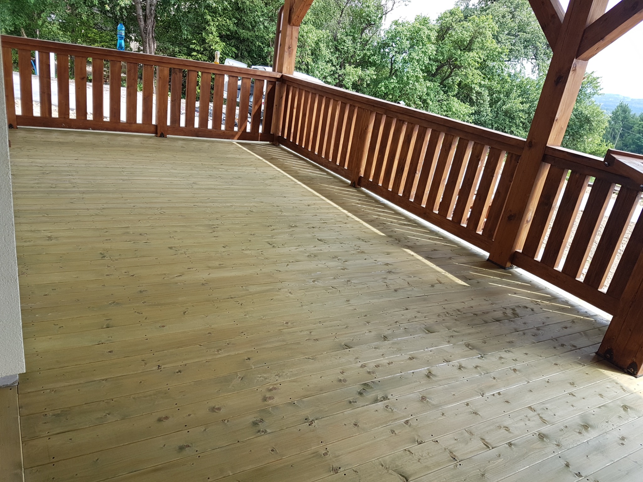 Świeżo wykończony taras z jasnego drewna z brązową balustradą, widok na zieleń w tle.