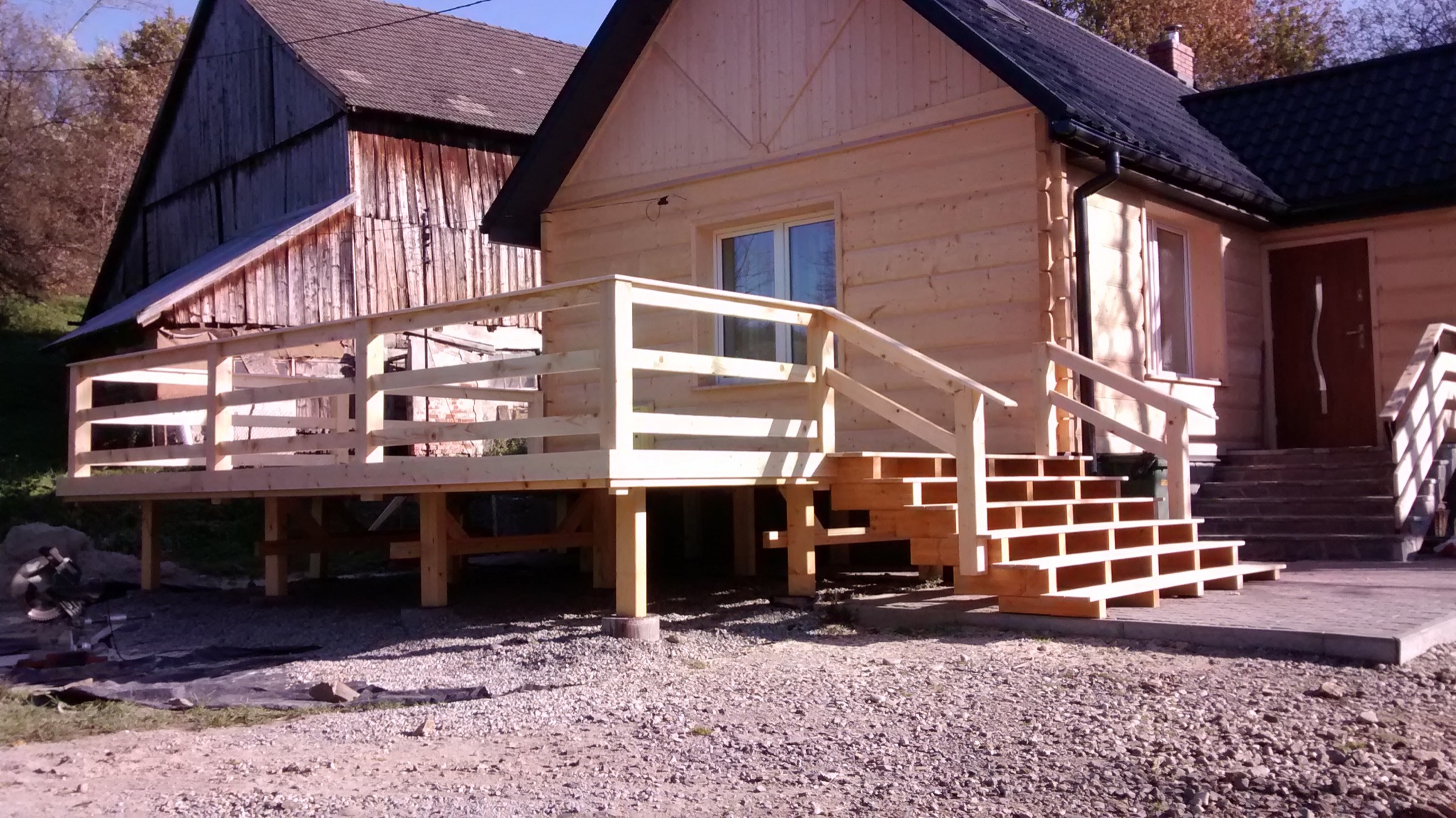 Surowy drewniany dom z nowo wybudowanym tarasem i schodami, widoczne ślady prac budowlanych na gruncie.
