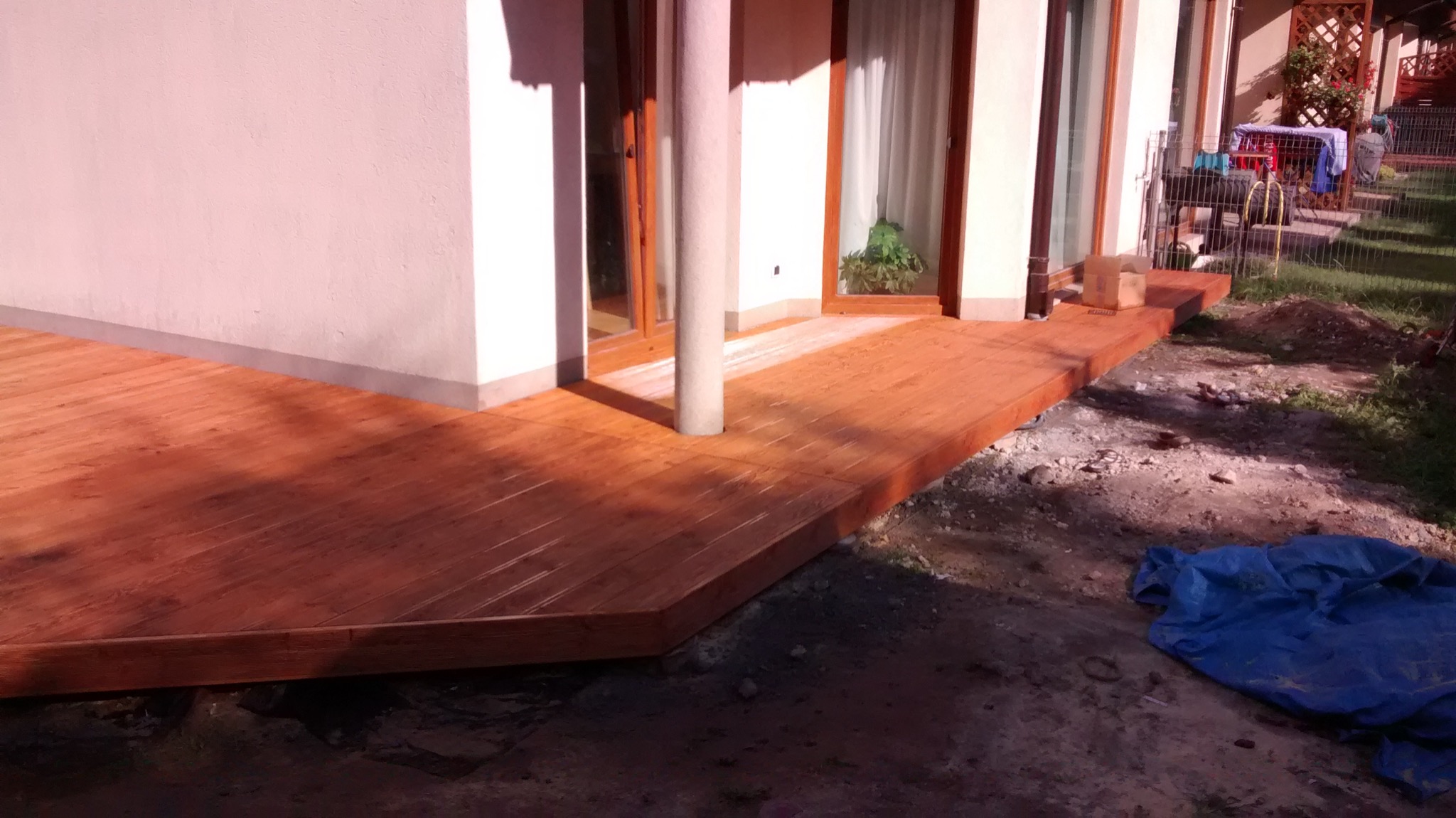 Nowo wybudowany taras z drewna egzotycznego, z kolumną wbudowaną w konstrukcję, widok na ogród i dom w tle.