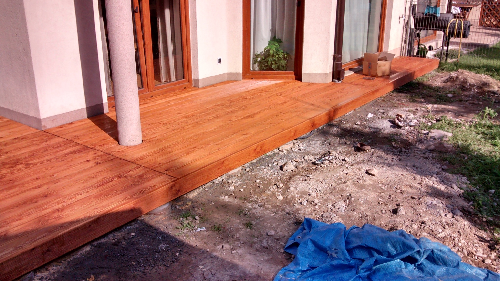 Nowy taras z desek kompozytowych w odcieniach brązu, z widocznym słupem podtrzymującym i fragmentem ogrodu w trakcie aranżacji.