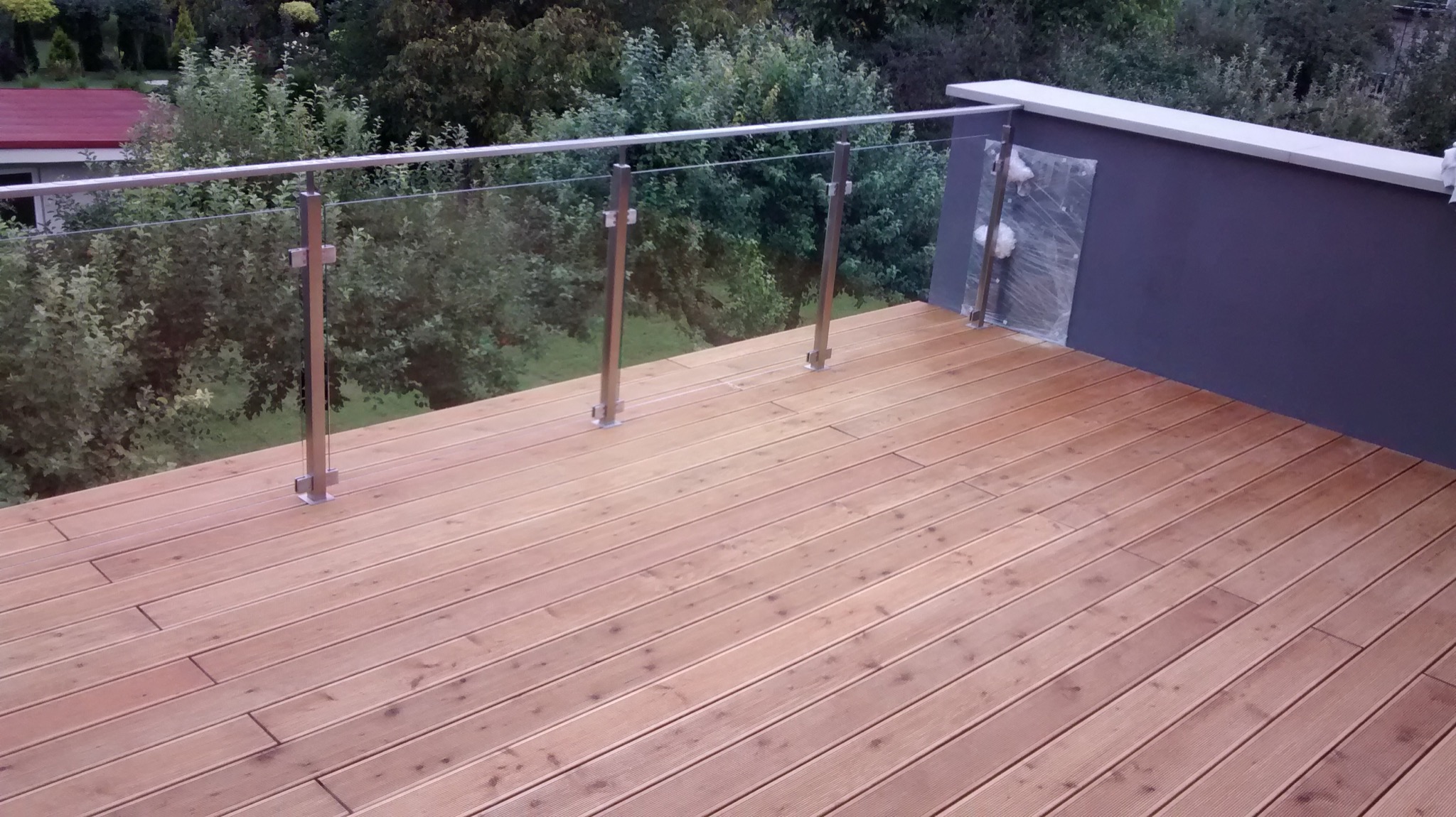 Nowoczesny taras z drewnianą podłogą i szklaną balustradą ze stalowymi słupkami, widok na zieleń w tle, folia ochronna na elemencie ściany.