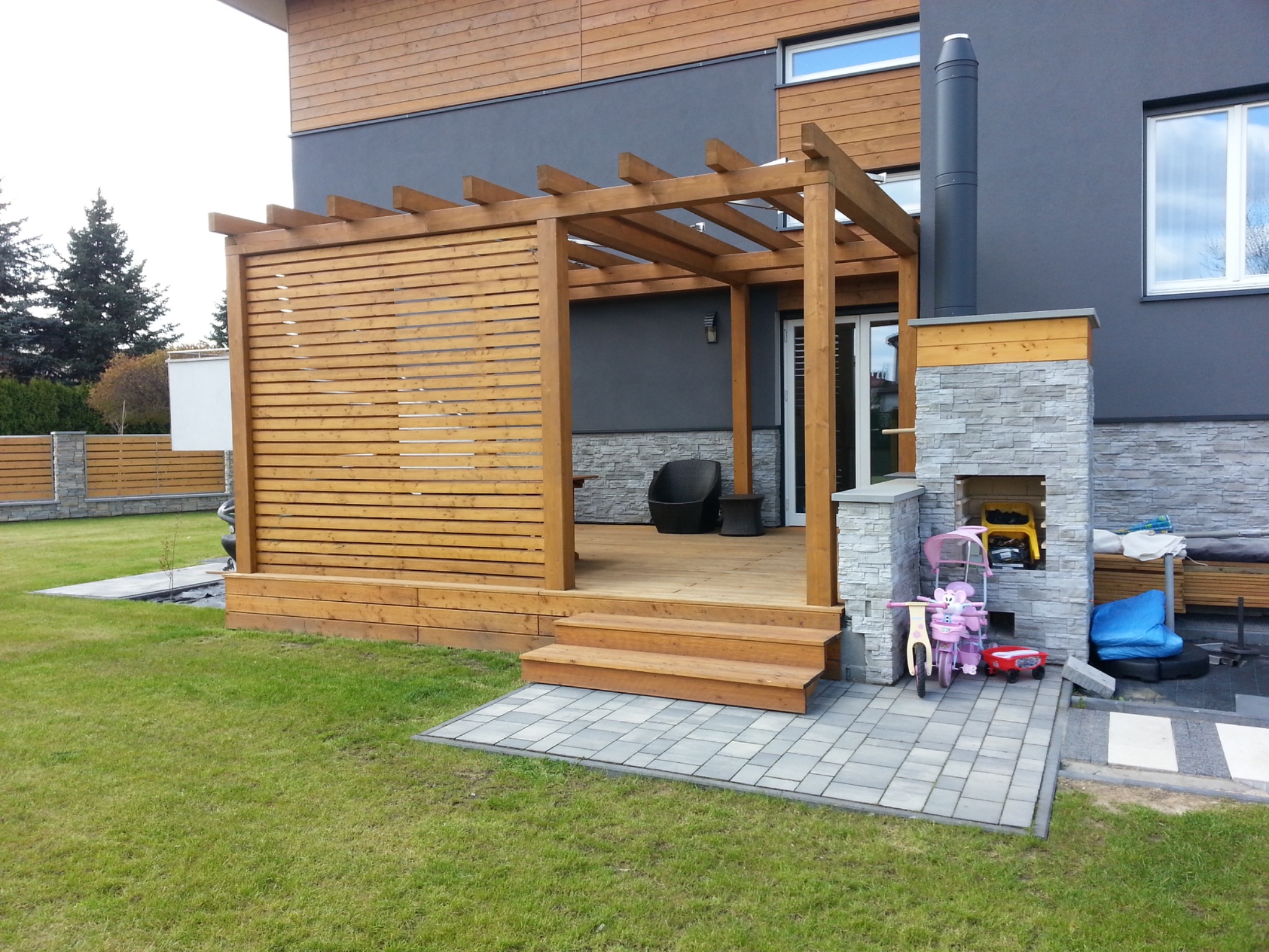 Drewniana pergola z ażurową ścianą boczną przylegająca do domu z kominkiem zewnętrznym, widok na taras z meblami ogrodowymi.