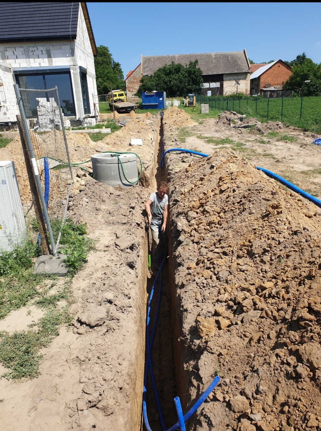 Pracownik kopie wąski wykop pod niebieskie rury instalacyjne obok betonowego kręgu studni, na tle budynek w stanie surowym otwartym i zabudowania gospodarcze.