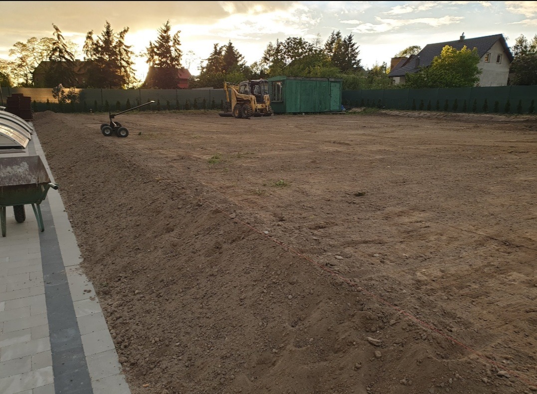 Przygotowany teren pod budowę z widocznym wykopem, wyrównaną ziemią, żółtą koparką w tle i zielonym barakiem, oświetlony popołudniowym słońcem.