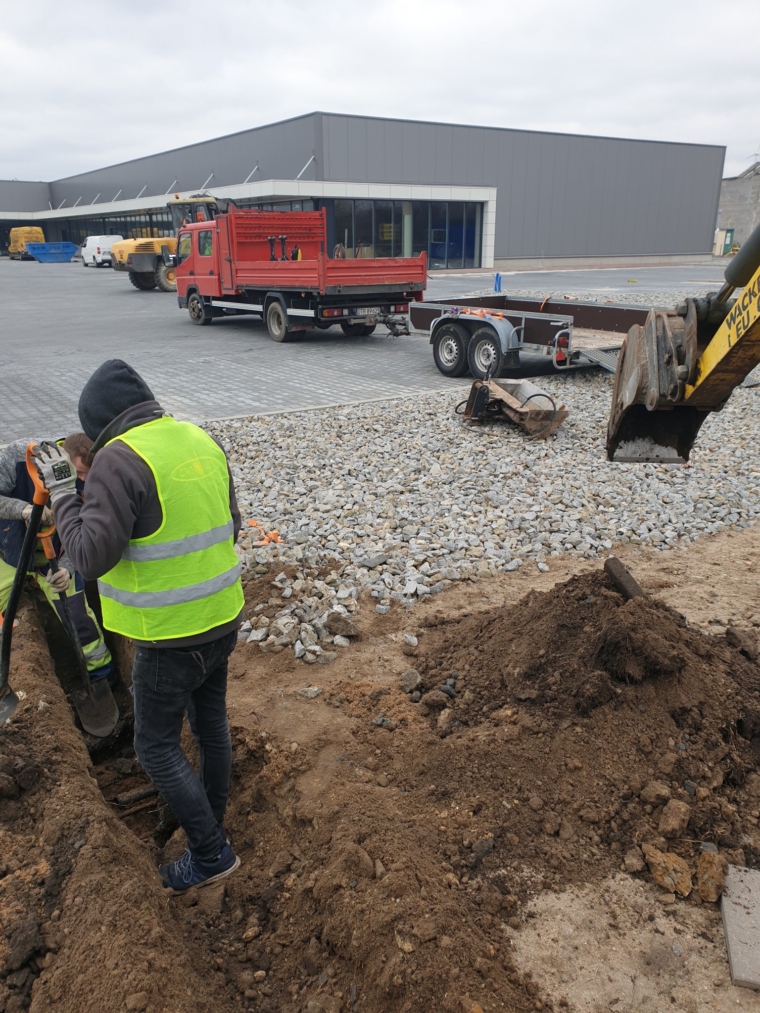 Dwóch pracowników w odblaskowych kamizelkach kopie wąski wykop obok stosu ziemi, w tle czerwona ciężarówka, przyczepa i koparka na placu budowy.