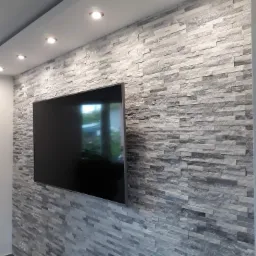 Ściana wyłożona szarymi płytkami imitującymi kamień, z zamontowanym telewizorem i oświetleniem punktowym w suficie.