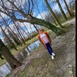 Kobieta w kurtce z pomarańczowym futerkiem stoi na tle parku z jeziorkiem, drzewami i błękitnym niebem z chmurami.