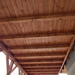 Part Home - Drewniana konstrukcja zadaszenia tarasu, widok od dołu, widoczne deski i belki, na zewnątrz kostka brukowa, samochód i osoba w tle.