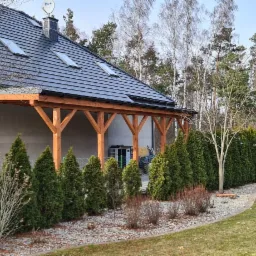 Nowoczesny dom jednorodzinny z ciemną dach&oacute;wką i drewnianą pergolą, widok na zadbany trawnik i rząd tuj w słoneczny dzień.