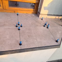 Part Home - Układanie płytek na balkonie z użyciem systemów poziomowania, widoczne okno i kwiaty w doniczce.