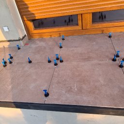 Part Home - Balkon w trakcie układania płytek, widoczne narzędzia poziomujące i krzyżyki dystansowe, w tle okno z drewnianą ramą i kwiaty w doniczce.