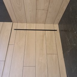 Part Home - Ułożone podłużne płytki podłogowe imitujące drewno, czarny odpływ liniowy przy ścianie, fragment marmurowej ściany i niebieski element instalacji w tle, widoczne buty osoby fotografującej.