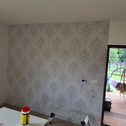 Part Home - Fragment pokoju po tapetowaniu ścian wzorzystą tapetą w odcieniach szarości, widoczna drabina, stół z narzędziami i otwarte drzwi balkonowe.