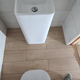 Part Home - Ukończona instalacja umywalki i toalety w nowoczesnej łazience z drewnopodobnymi płytkami podłogowymi, widok z góry na stopy instalatora w ubrudzonych farbą butach i spodniach roboczych...