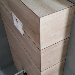 Part Home - Obudowa stelaża podtynkowego WC wykończona płytkami imitującymi drewno, widoczne wystające rurki instalacyjne i otwór rewizyjny z fragmentami folii ochronnej, w tle szara ściana i podłoga.