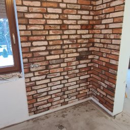 Part Home - Fragment ściany wewnętrznej wyłożonej dekoracyjną cegłą w odcieniach czerwieni i bieli, z widocznymi gniazdkami elektrycznymi, surowy beton na podłodze, okno z drewnianą ramą po lewej stronie...