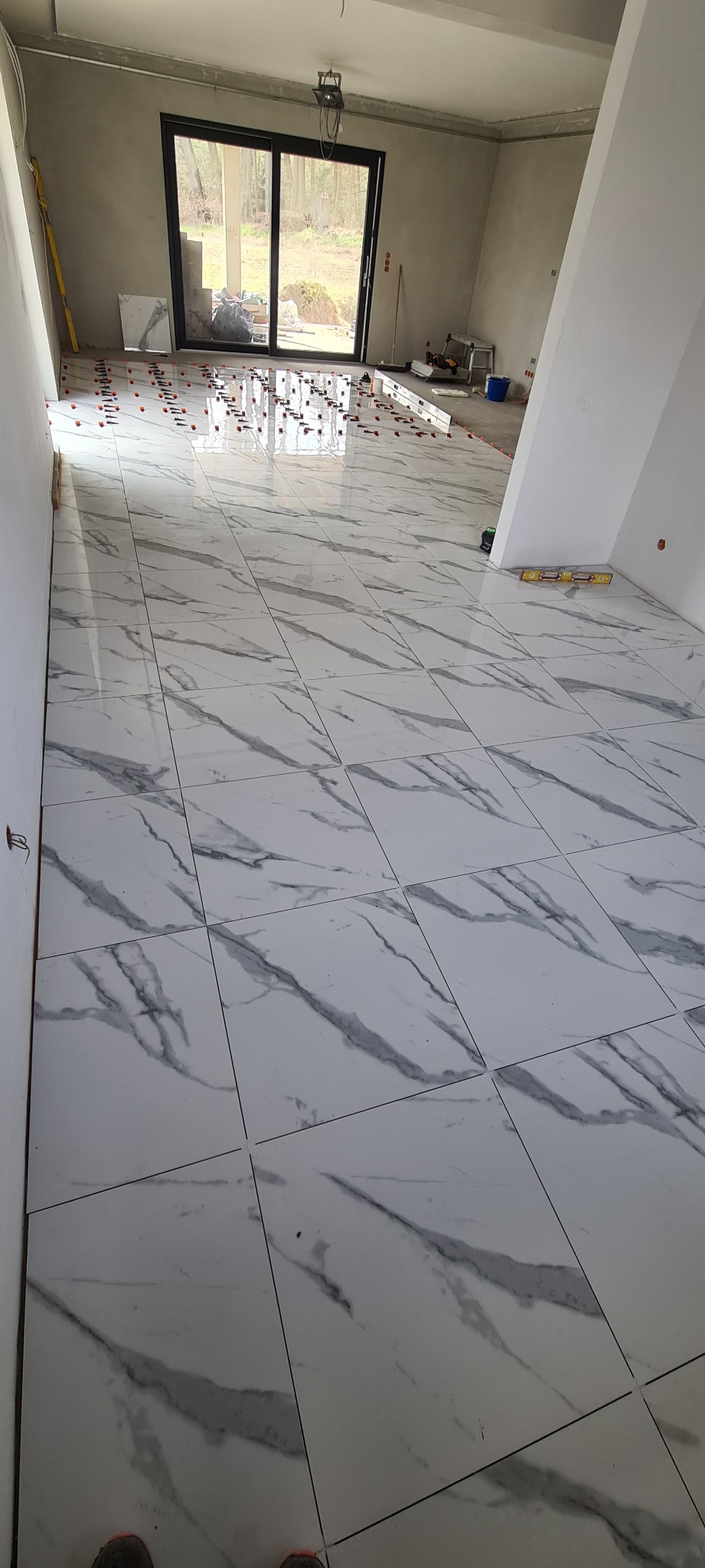 Układanie białych płytek podłogowych imitujących marmur w jasnym pomieszczeniu z dużym oknem balkonowym. Widoczne narzędzia glazurnicze i system poziomowania płytek.