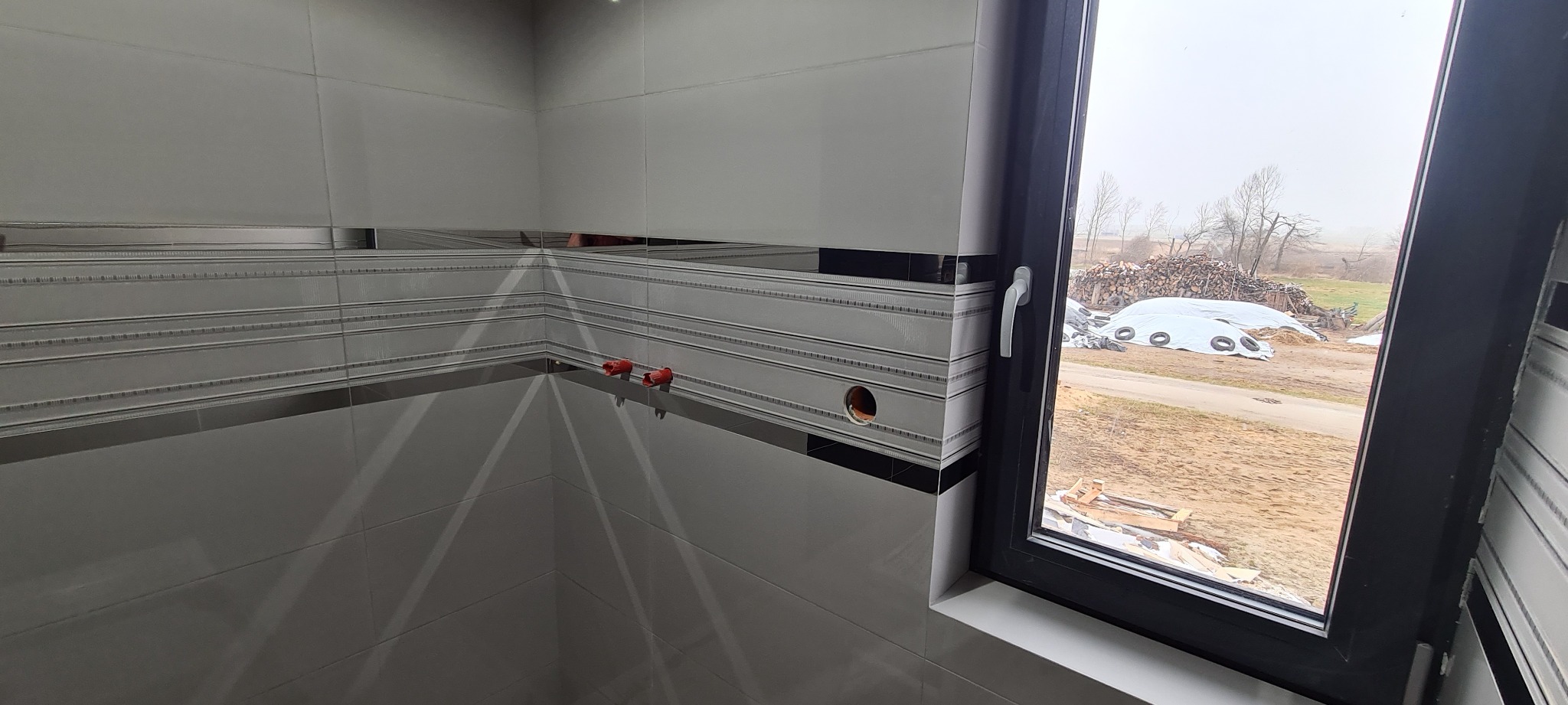 Szare płytki z dekoracyjnymi paskami w nowo wykończonej łazience, widoczne wystające rurki i otwór w ścianie, okno z widokiem na stos drewna opałowego.