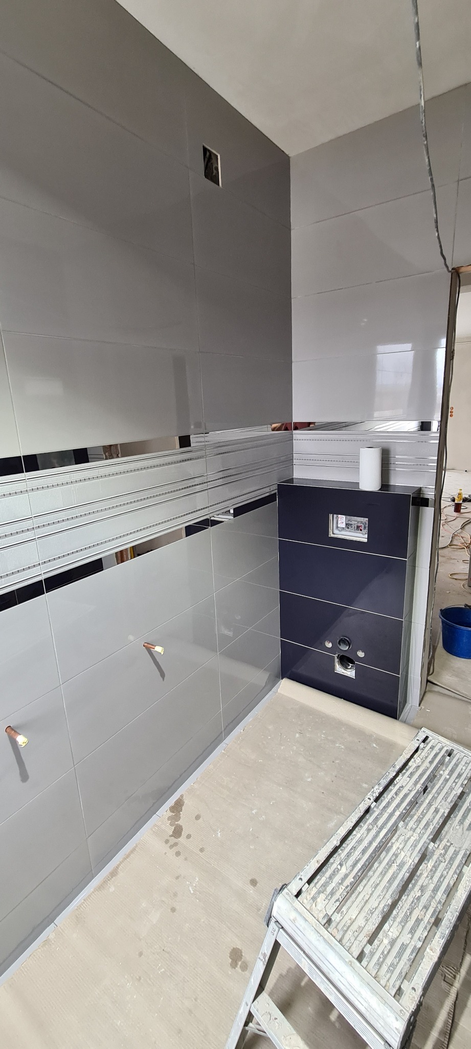 Szare i białe płytki ścienne z dekoracyjnymi pasami w łazience w trakcie remontu, widoczne wystające rury i zabudowa toalety podtynkowej z ciemnymi płytkami, drabina malarska w dolnym rogu.