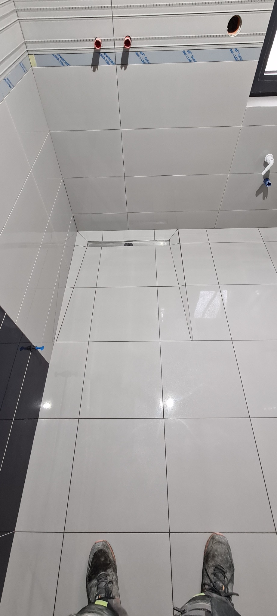 Nowoczesna łazienka w trakcie wykańczania, widok z góry na jasne, błyszczące płytki ceramiczne na ścianach i podłodze, z widocznym odpływem liniowym, wystającymi rurami z sufitu i butami osoby...