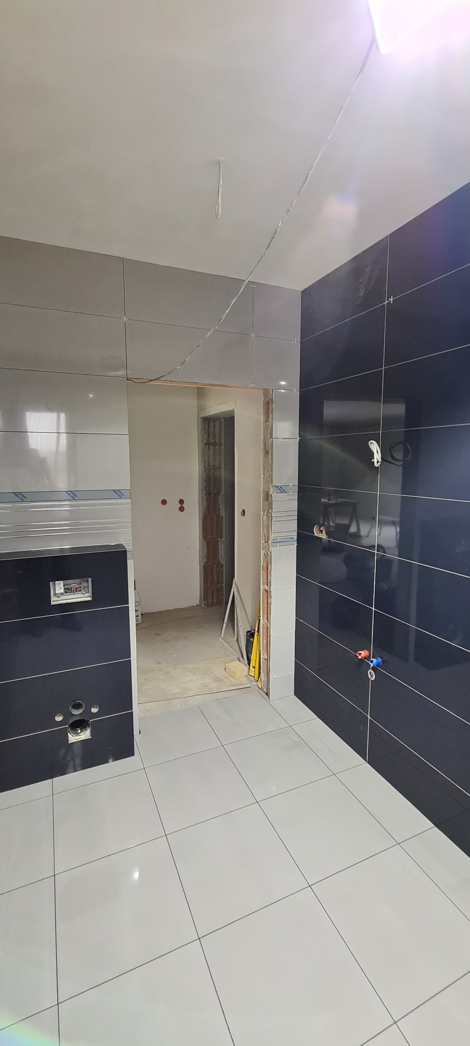 Wnętrze łazienki w trakcie remontu: ułożone płytki na ścianach w kolorach szarości i granatu, biała podłoga z płytek, widoczne przewody i niezakończone prace przy drzwiach.