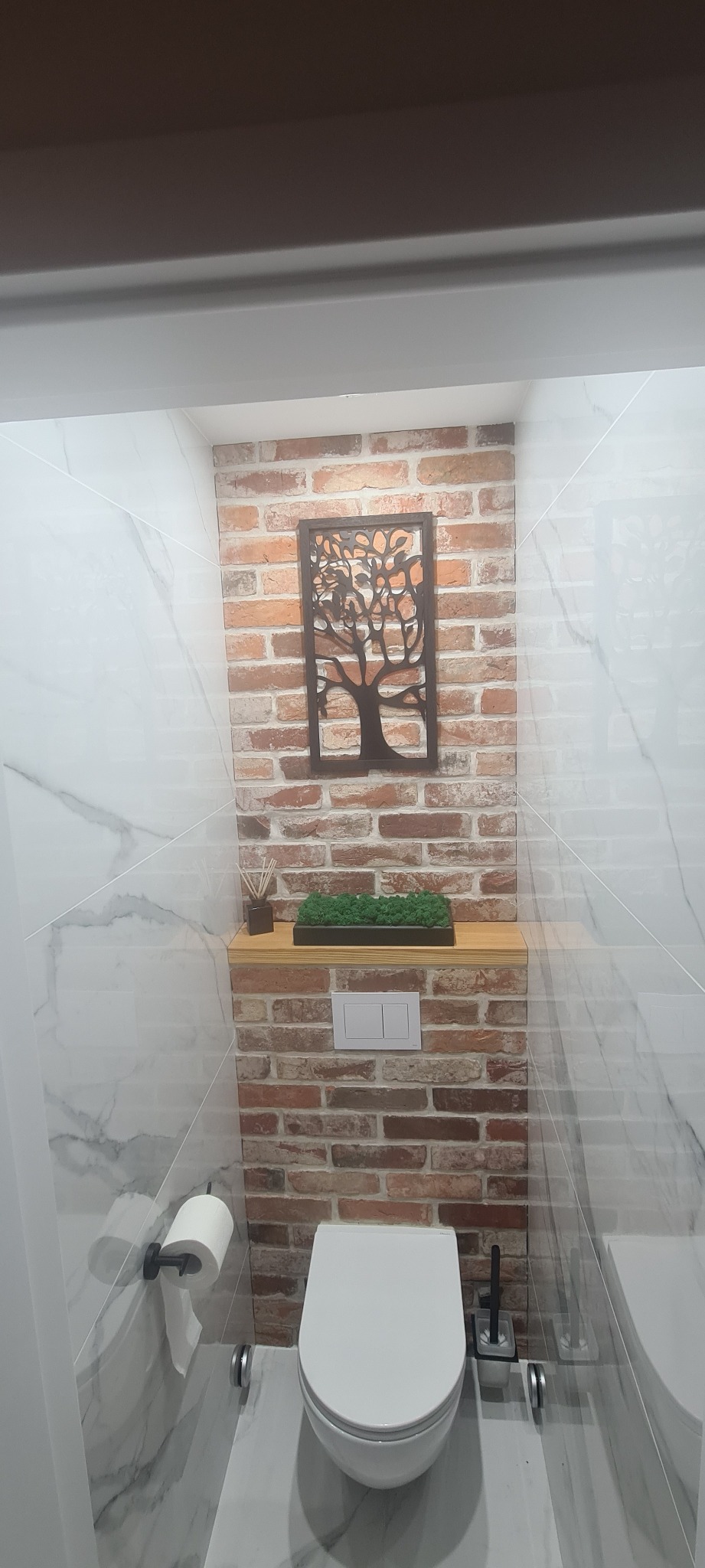 Wnętrze łazienki z toaletą wiszącą, obudowaną cegłą klinkierową, dekoracją ścienną w kształcie drzewa, półką z mchem i marmurowymi płytkami na ścianach.