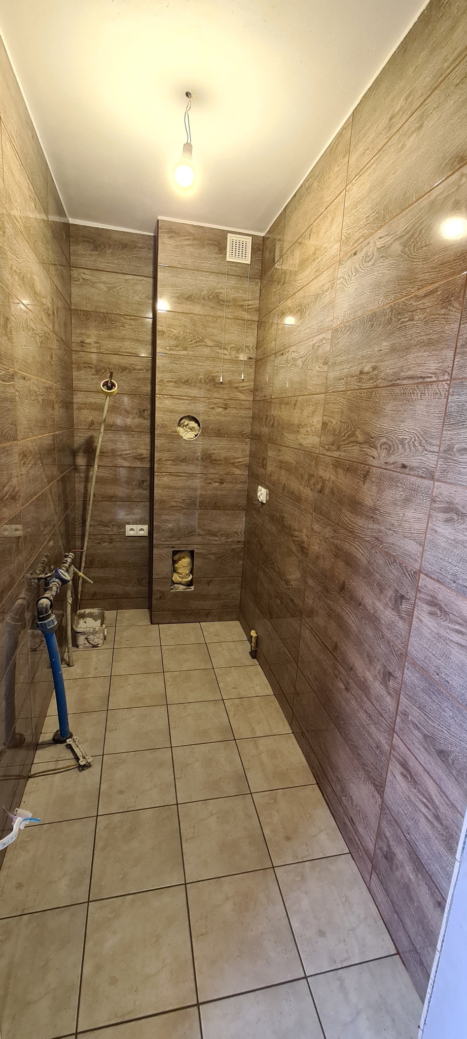 Wnętrze łazienki w trakcie remontu, z brązowymi płytkami imitującymi drewno na ścianach i beżowymi płytkami na podłodze, widoczne instalacje hydrauliczne, otwory w ścianach i wisząca żarówka.