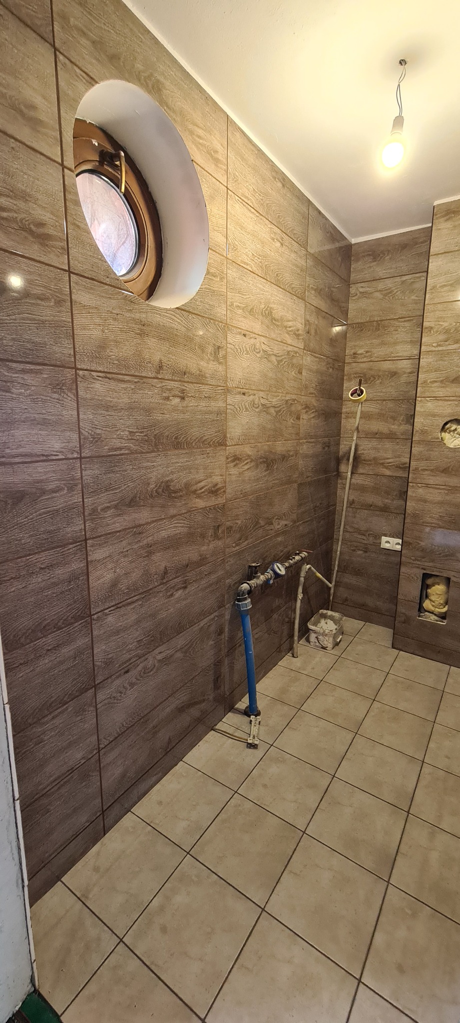 Wykończona łazienka z brązowymi płytkami imitującymi drewno na ścianach, okrągłe okno w górnej części ściany, widoczne rury i instalacje wodne przed zabudową, beżowa podłoga z płytek.