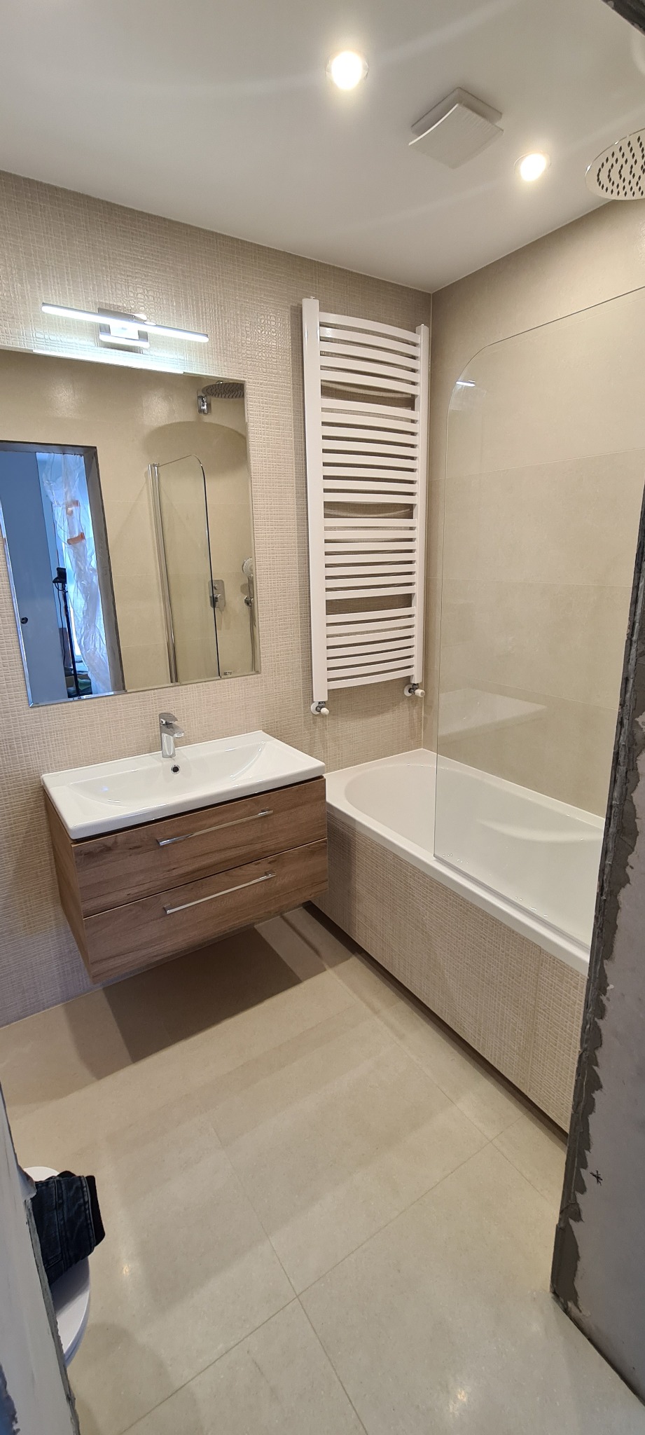 Wykończona łazienka w beżowych odcieniach z wiszącą szafką z umywalką z drewnopodobnymi szufladami, grzejnikiem drabinkowym i wanną z parawanem nawannowym, widok na lustro z oświetleniem i fragment...