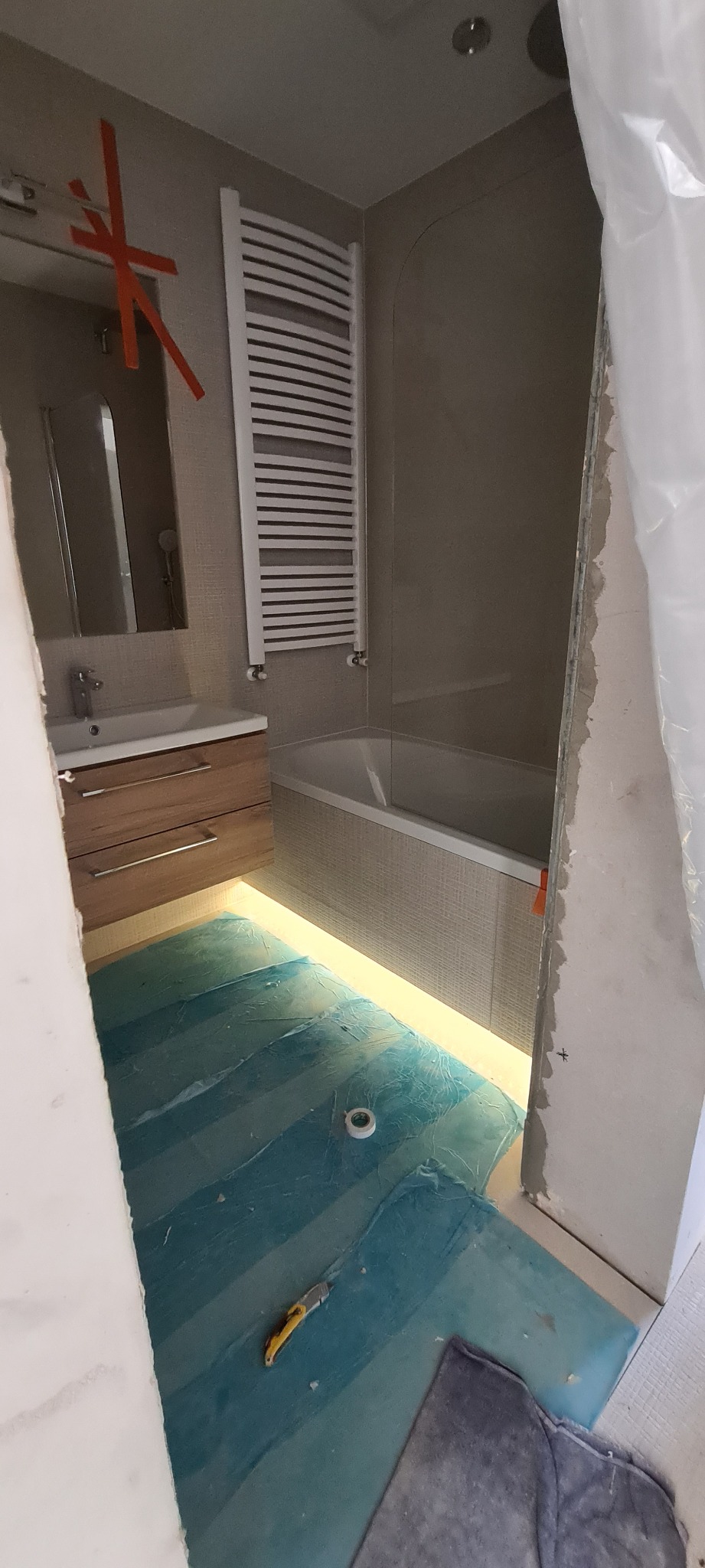 Łazienka w trakcie remontu: zabezpieczona folią podłoga z nożykiem, umywalka z szafką, grzejnik drabinkowy i wanna z obudową, ściany częściowo wykończone płytkami, widoczne ślady prac budowlanych.