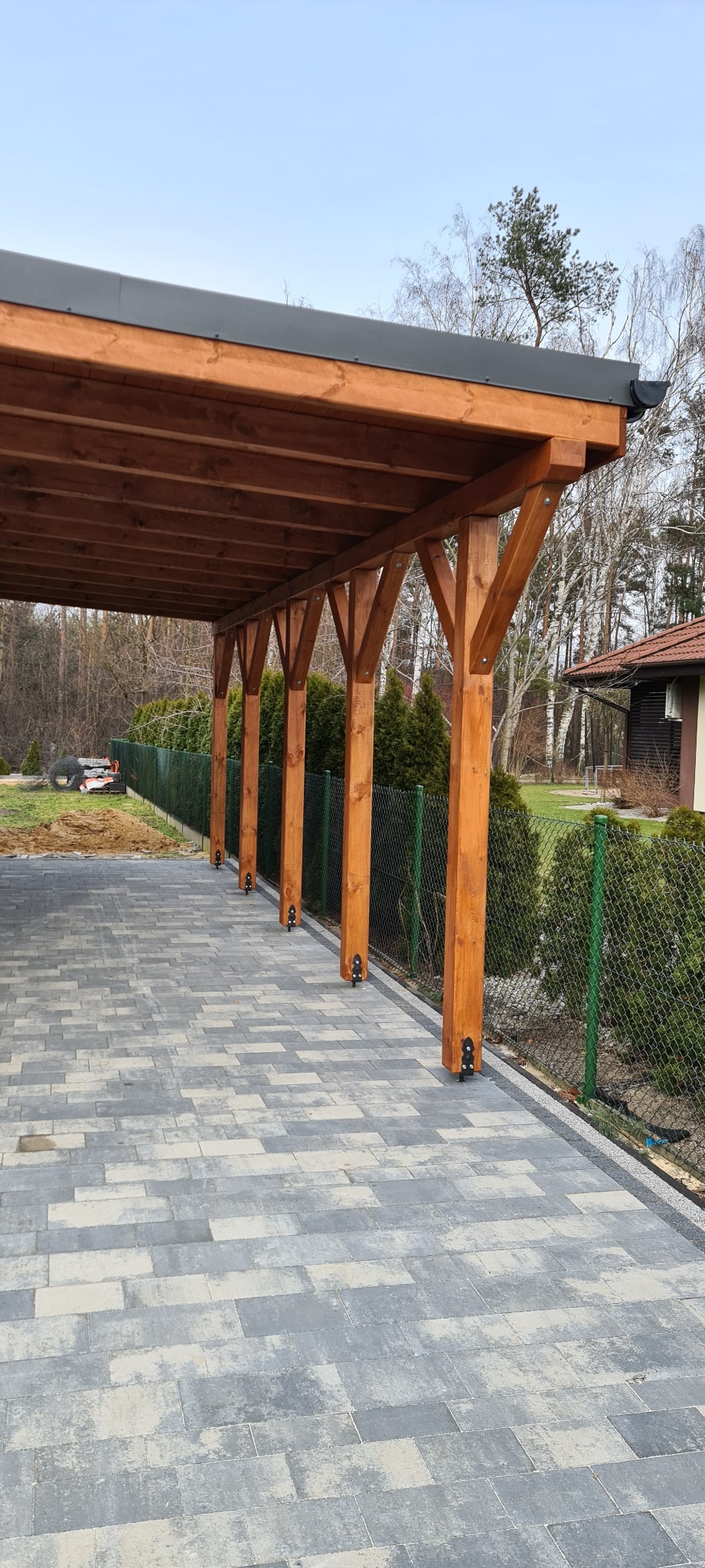 Zadaszona wiata garażowa z drewnianą konstrukcją, szara blacha na dachu, podłoże z kostki brukowej, widok z boku na tle zieleni i domu.