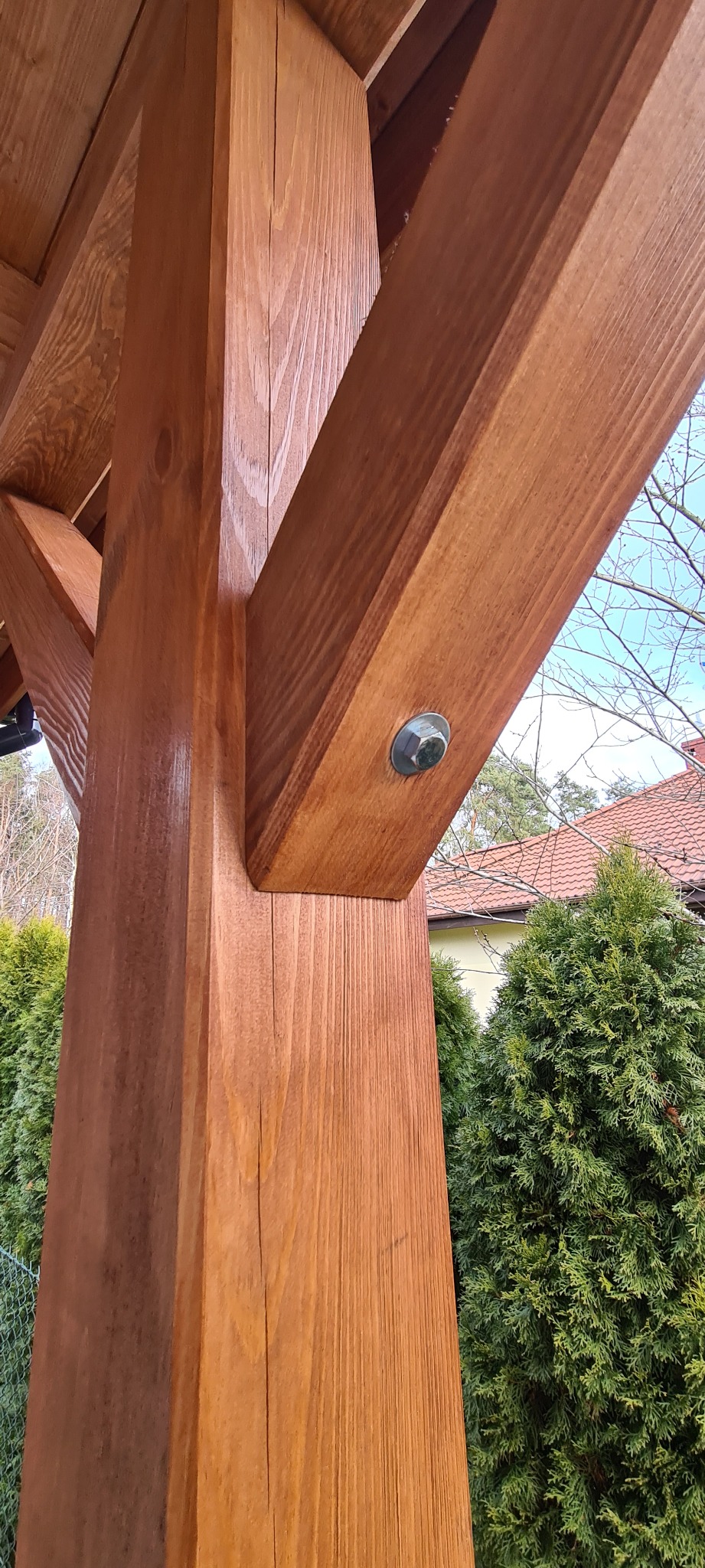 Połączenie drewnianych belek konstrukcyjnych altany za pomocą śruby z podkładką, widoczne usłojenie drewna i elementy otoczenia.