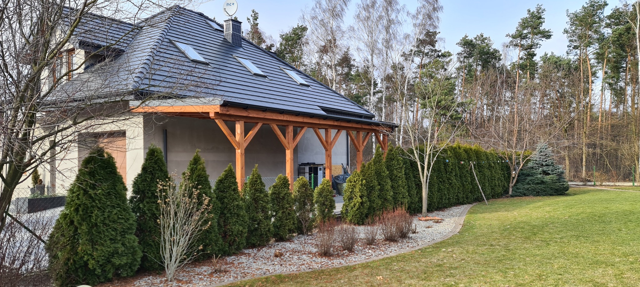 Nowoczesny dom jednorodzinny z ciemną dachówką i drewnianą pergolą, widok na zadbany trawnik i rząd tuj w słoneczny dzień.