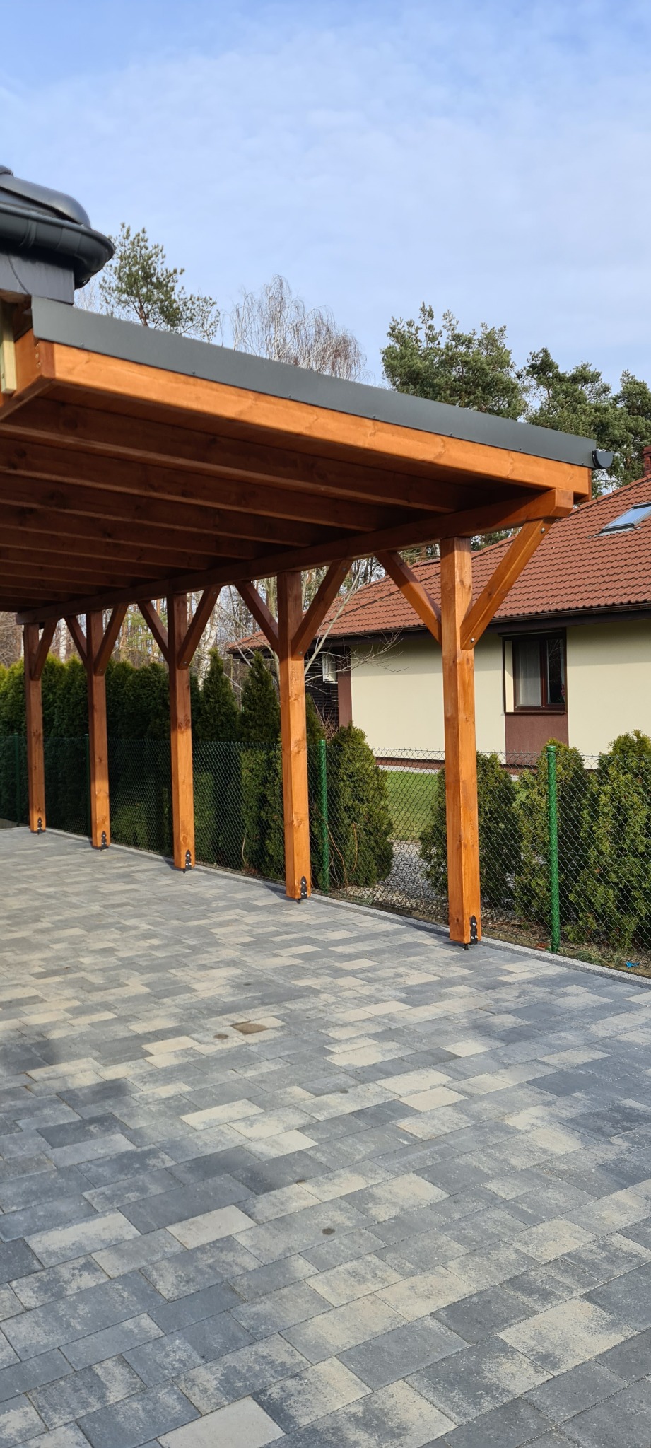 Drewniana wiata garażowa z płaskim, grafitowym dachem, wsparta na sześciu słupach; podłoże wyłożone szarą kostką brukową; w tle dom jednorodzinny z brązową dachówką.