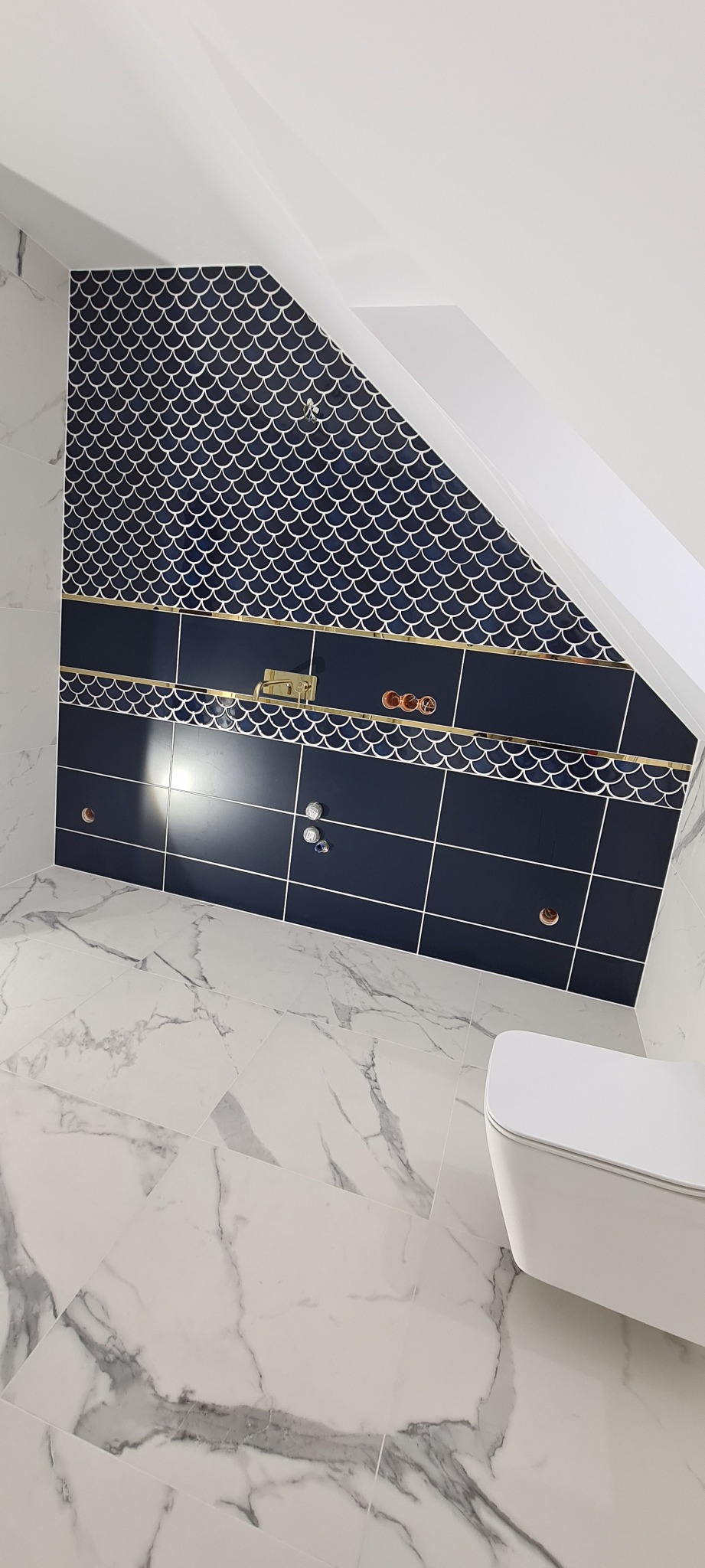 Wykończona łazienka na poddaszu z niebieskimi płytkami w kształcie rybich łusek, białymi marmurowymi płytkami podłogowymi i wiszącą toaletą.