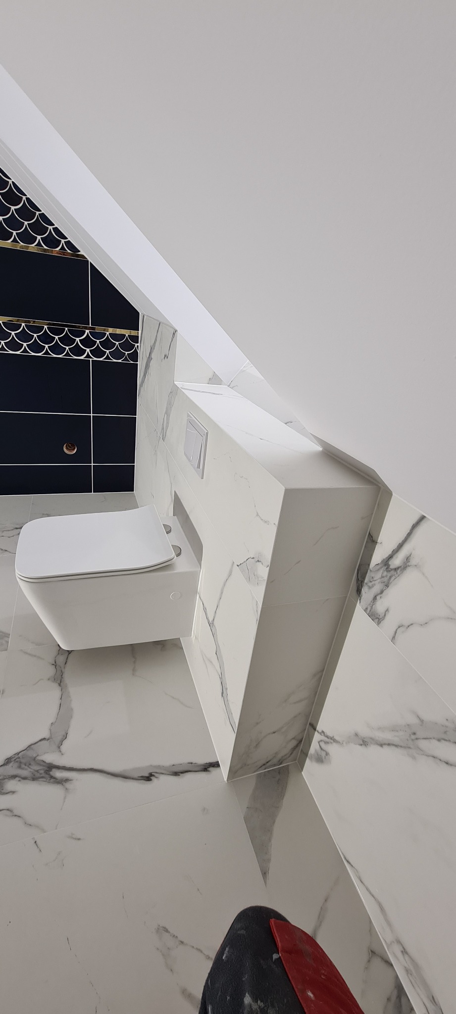 Nowoczesna toaleta podwieszana z zabudową w marmurkowe płytki, czarne płytki z motywem rybich łusek w górnej części ściany, biały sufit pod skosem.